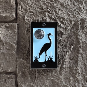 May include: A black doorbell button with a blue background and a silhouette of a heron standing in water. The button has a silver doorbell button.