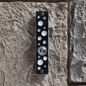 May include: A black doorbell with a silver button and a pattern of white circles. The doorbell is mounted on a stone wall.