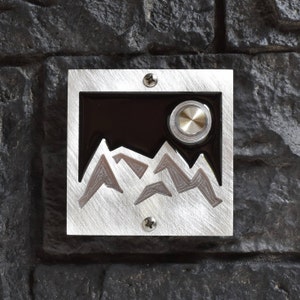 May include: A silver doorbell with a black background and a mountain range design. The doorbell button is round and silver.