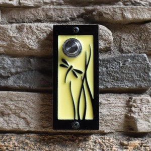 Peut inclure: Un bouton de sonnette noir avec un fond jaune et un motif de libellule. Le bouton est monté sur un mur en pierre.