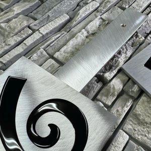 May include: A brushed silver metal house number plaque with a black swirl design. The number 6 is visible in the swirl.