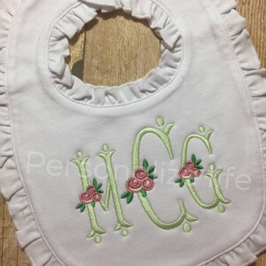 May include: White baby bib with a ruffled edge and a green embroidered monogram with pink flowers. The monogram is "MCC".