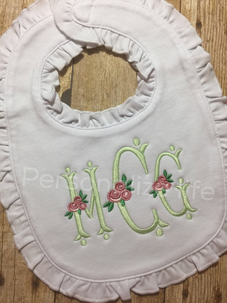 May include: White baby bib with a ruffled edge and a green embroidered monogram with pink flowers. The monogram is "MCC".