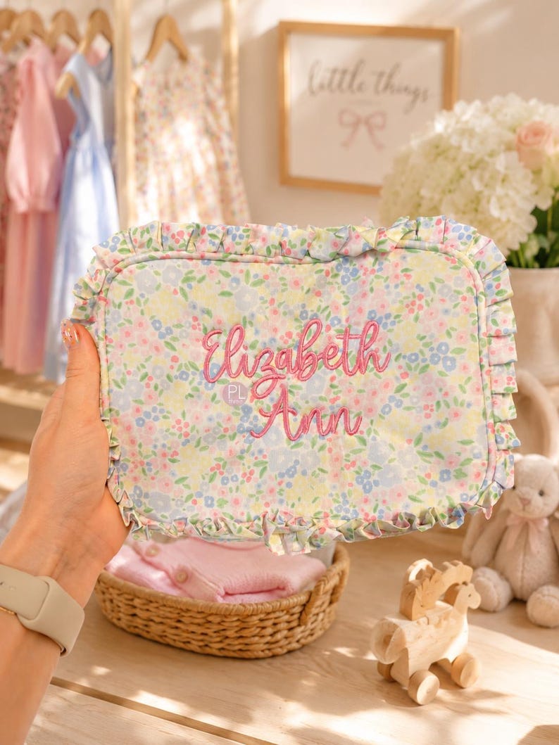 May include: A floral-patterned, rectangular pillow with ruffled edges. The pillow features the embroidered name "Elizabeth Ann" in pink thread. The background includes a wicker basket with pink clothing, a wooden toy, and a framed sign.