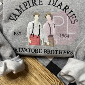 May include: A gray sweatshirt with the words "VAMPIRE DIARIES" arched across the top in black embroidery. Below the text are two figures and the words "SALVATORE BROTHERS". The sweatshirt also features the text "EST. 1864".