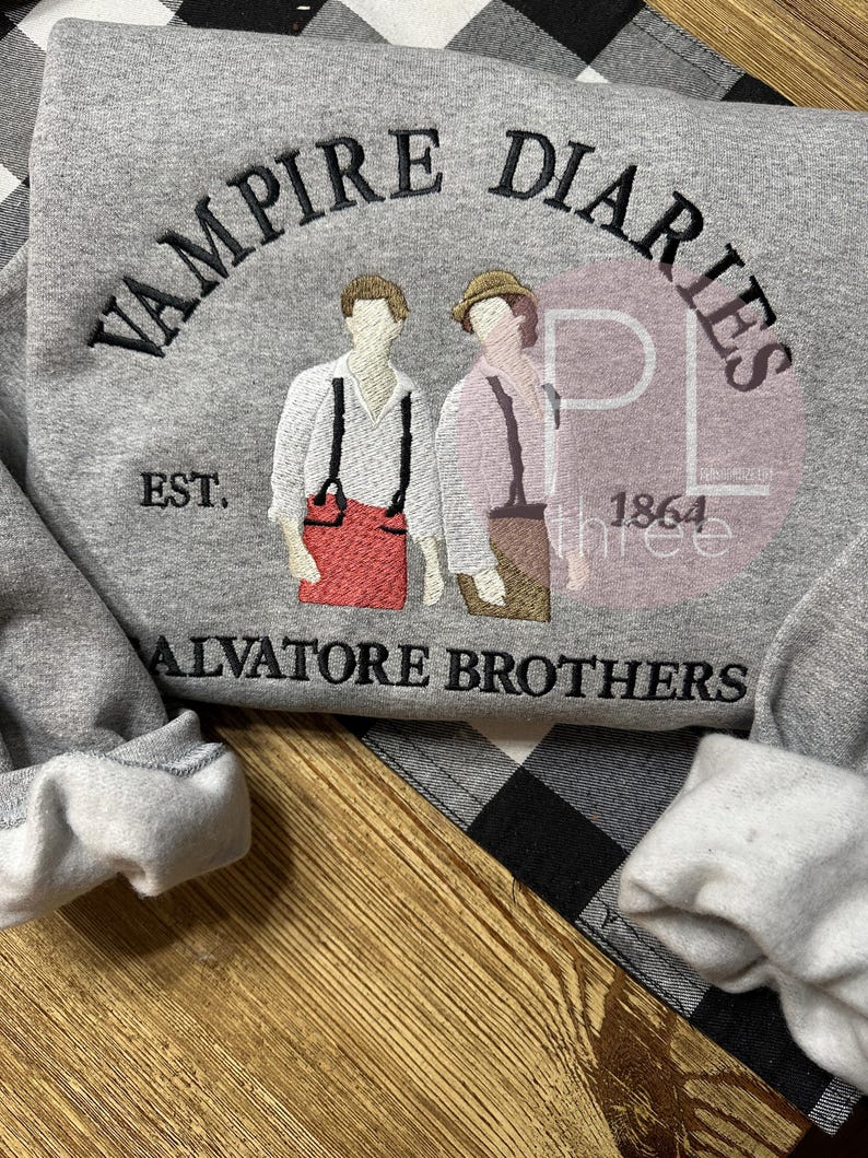 May include: A gray sweatshirt with the words "VAMPIRE DIARIES" arched across the top in black embroidery. Below the text are two figures and the words "SALVATORE BROTHERS". The sweatshirt also features the text "EST. 1864".