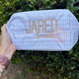 May include: A rectangular, beige and white gingham patterned bag with the name "JARED" embroidered in a light brown thread. The name "brooks" is embroidered below. The bag is held against a backdrop of green foliage.