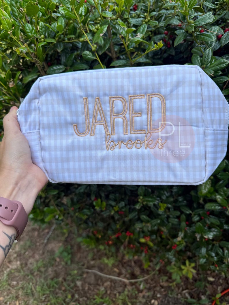 May include: A rectangular, beige and white gingham patterned bag with the name "JARED" embroidered in a light brown thread. The name "brooks" is embroidered below. The bag is held against a backdrop of green foliage.