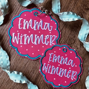 May include: Two pink and white polka dot fabric name tags with a turquoise border. The tags are shaped like a cloud and have the name "Emma Wimmer" embroidered in white on them. The tags are attached to a light blue ribbon.
