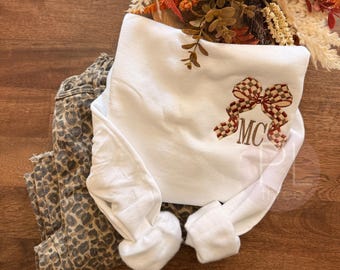 May include: A white long-sleeve sweatshirt featuring an embroidered brown and tan checkered bow design above the monogrammed letters MC. The sweatshirt is paired with a pair of brown and black leopard print denim pants.
