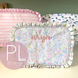 May include: Three rectangular, ruffled pouches. One is pink gingham, another has a floral pattern with the name "Arya" embroidered, and the third features a duck pattern. The pouches are likely for storage or organization.