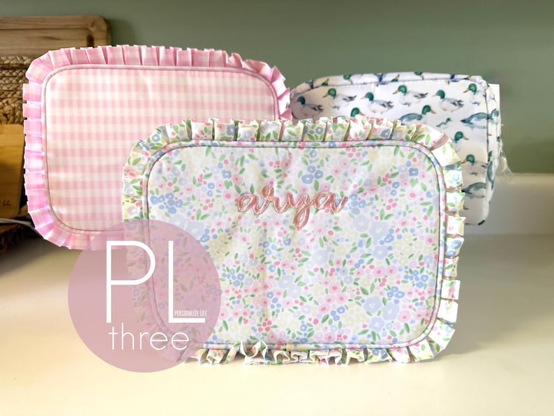 May include: Three rectangular fabric pouches with ruffled edges. One is pink gingham, another features a floral pattern with the name "Arya" embroidered, and the third has a duck pattern. The pouches are likely for storage or organization.