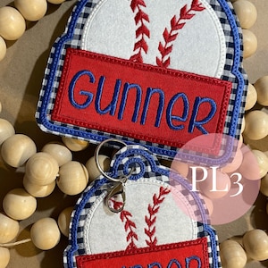 May include: Two baseball-themed keychains with a blue and white gingham pattern. The keychains feature a white baseball with red stitching and a red banner with the name "Gunner" in blue lettering. The larger keychain has a silver key ring.