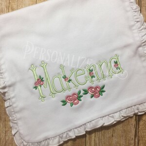 May include: White baby blanket with a ruffled edge and the name "Makenna" embroidered in green with pink roses.