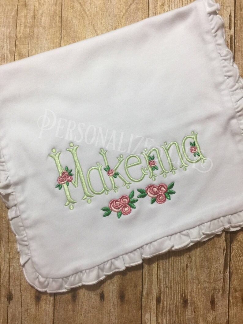 May include: White baby blanket with a ruffled edge and the name "Makenna" embroidered in green with pink roses.