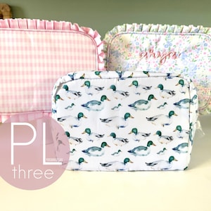 May include: Three cosmetic bags with different patterns. One is pink gingham, another has a floral print with the name "Arya" embroidered, and the third features a duck pattern. The bags have ruffled edges.