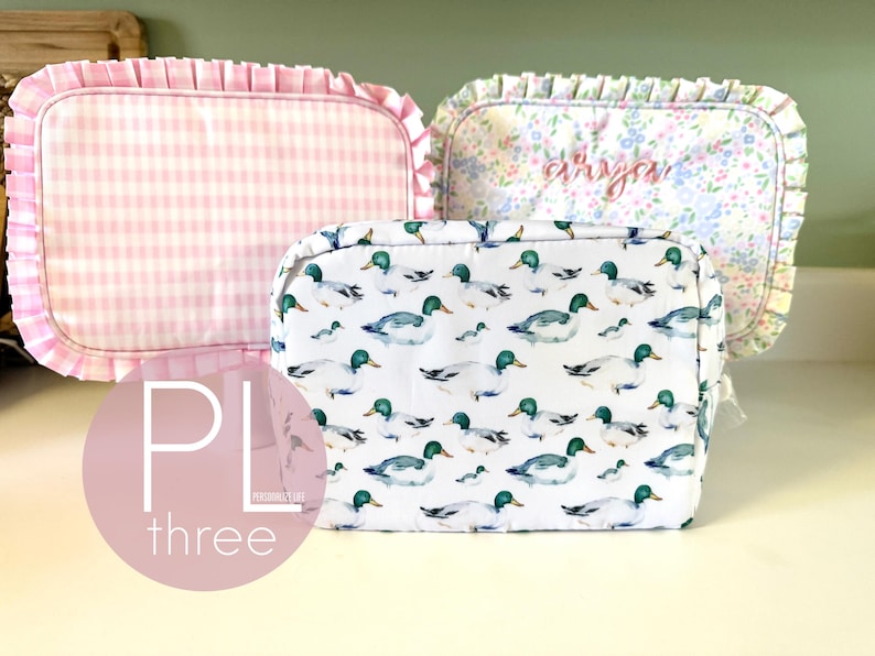 May include: Three cosmetic bags with different patterns. One is pink gingham, another has a floral print with the name "Arya" embroidered, and the third features a duck pattern. The bags have ruffled edges.