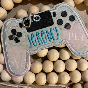 May include: A gray and pink felt video game controller keychain with the name "Jeremy" and the text "PL3" embroidered on it.