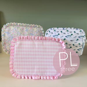 May include: Three rectangular cosmetic bags with ruffled edges. One bag has a pink gingham pattern, another has a floral pattern, and the third features a duck pattern. The bags are displayed on a white surface.