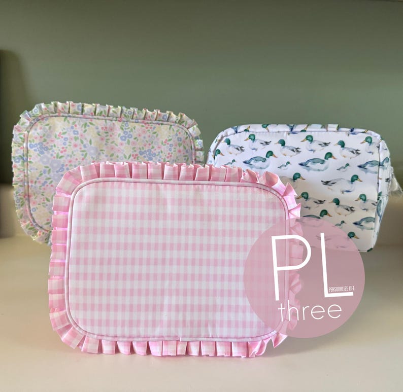 May include: Three rectangular cosmetic bags with ruffled edges. One bag has a pink gingham pattern, another has a floral pattern, and the third features a duck pattern. The bags are displayed on a white surface.