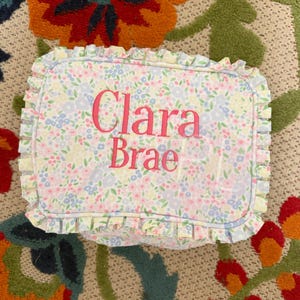 May include: Rectangular floral print pillow with the name "Clara Brae" embroidered in pink. The pillow has a ruffled edge and a pattern of small blue, pink, and yellow flowers.