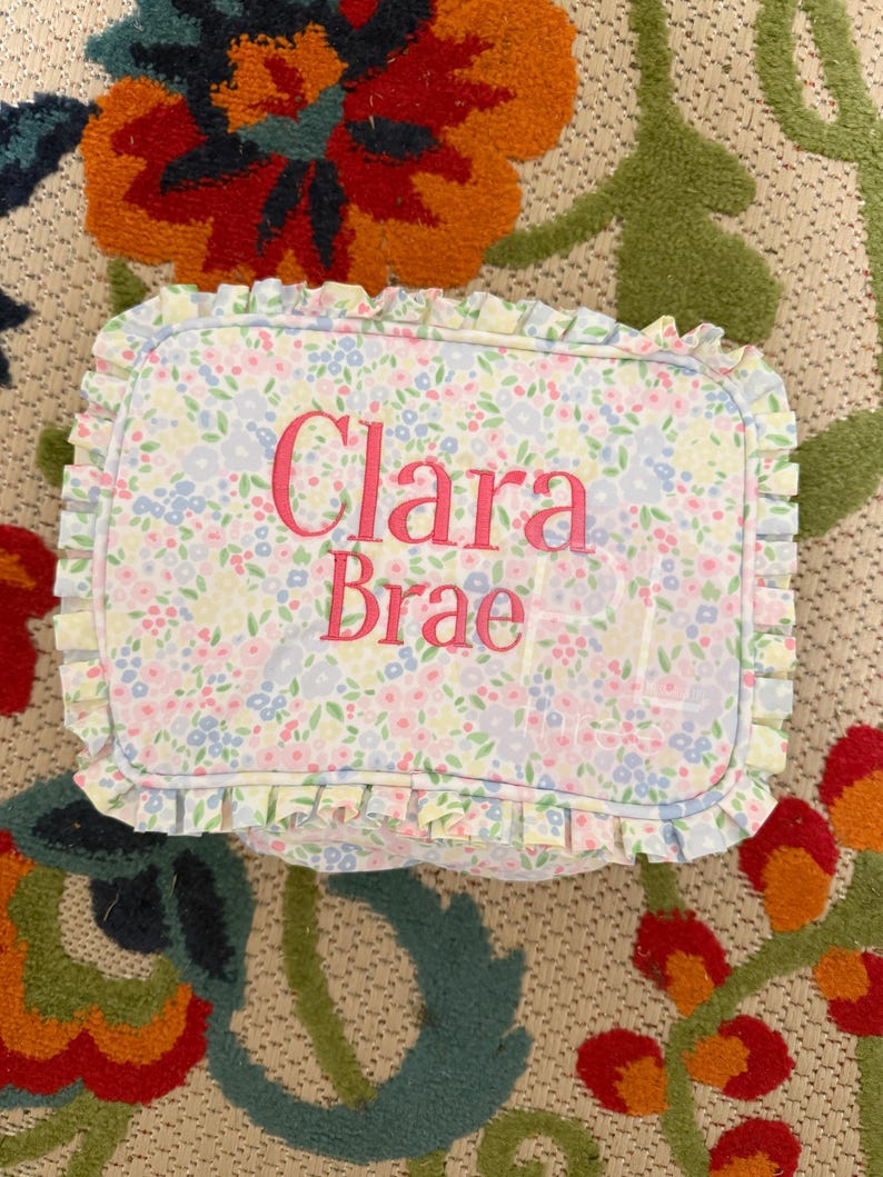 May include: Rectangular floral print pillow with the name "Clara Brae" embroidered in pink. The pillow has a ruffled edge and a pattern of small blue, pink, and yellow flowers.