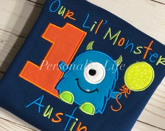May include: A blue t-shirt with the text "Our Lil' Monster" in green and orange. The number "1" is orange with an orange outline. A blue monster with one eye and a toothy grin holds a yellow balloon with green polka dots. The name "Austin" is written in orange and green below the monster.