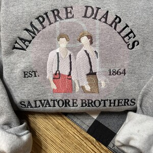 May include: A gray sweatshirt featuring the text "VAMPIRE DIARIES" arched above an illustration of two figures. Below the illustration are the words "SALVATORE BROTHERS" and the year "1864". The sweatshirt is on a black and white checkered surface.
