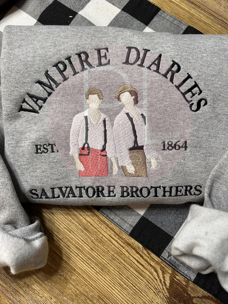 May include: A gray sweatshirt featuring the text "VAMPIRE DIARIES" arched above an illustration of two figures. Below the illustration are the words "SALVATORE BROTHERS" and the year "1864". The sweatshirt is on a black and white checkered surface.