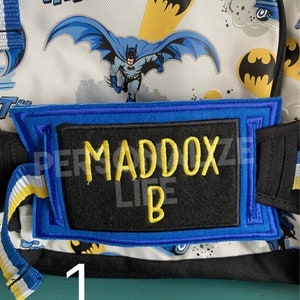 May include: A blue and black personalized name tag with yellow stitching. The tag reads "Maddox B" and is attached to a white and blue Batman themed backpack.