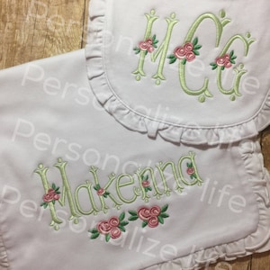 May include: White bib and burp cloth with green embroidered lettering and pink floral accents. The bib has a ruffled edge. The burp cloth has the name "Makenna" embroidered on it.
