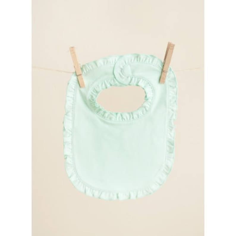 May include: A light green baby bib with a ruffled edge. The bib has a small opening at the top for the neck.