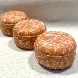 May include: Three round, solid shampoo bars in a light terracotta color with visible white flecks throughout the surface. Each bar has a cylindrical shape with a flat top and bottom, arranged in a diagonal line from the foreground to the back.