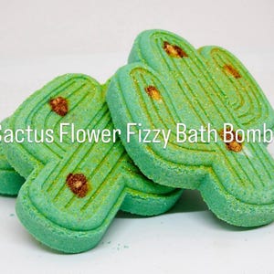 May include: Two light green cactus-shaped bath bombs with textured surfaces, gold glitter accents, and brown detailing. The text Cactus Flower Fizzy Bath Bombs is overlaid on the image. These items have a flat, molded design with vertical lines.