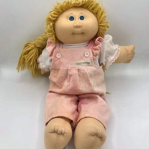 Puede incluir: Una muñeca Cabbage Patch Kids con cabello de lana rubia peinado en una trenza lateral, ojos azules y cara redonda. La muñeca viste una camisa blanca de manga corta con ribete de encaje y un overol de pana rosa con dos botones blancos y etiqueta.