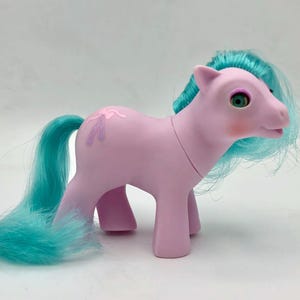 Puede incluir: Un poni de juguete rosa vintage con pelo y cola turquesa. El poni tiene un diseño de zapatilla de ballet en un lado y ojos verdes. El poni está sobre una superficie blanca.