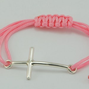 May include: A pink braided cord bracelet with a silver cross charm. The cross is a simple design with a curved bar.