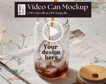 May include: A clear glass can-shaped mockup with ice and a beverage being poured into it. The glass has the text "Your design here" and a play button overlay. The image includes the text "Video Can Mockup" and "1 MP4 video file & 1 JPG image file".