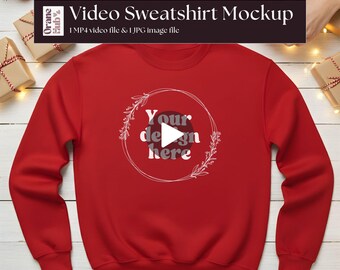 May include: A red sweatshirt with a circular design that says "Your design here". The sweatshirt is displayed on a white wooden surface, with Christmas decorations in the background. The text "Video Sweatshirt Mockup" is at the top.
