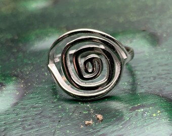 May include: A silver-colored ring with a spiral design. The ring features a central, tightly wound coil surrounded by a larger, open spiral. The ring is set against a green and black textured surface.
