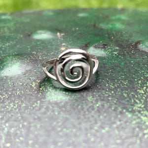 May include: A silver-colored ring with a spiral design. The ring features a coiled center, resembling a rose or a nautilus shell. The ring is set against a green and brown background.