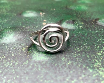 May include: A silver-colored ring with a spiral design. The ring features a coiled center, resembling a rose or a nautilus shell. The ring is set against a green and brown background.