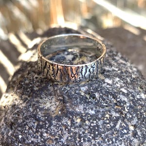 May include: A silver ring with a textured bark-like pattern. The ring is set on a dark gray stone, with a blurred background. The ring is a band style, with a detailed design around the entire circumference.