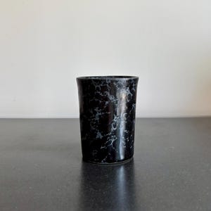 May include: A black ceramic tumbler with a marble-like pattern of white and gray veining. The cylindrical cup has a smooth, glossy finish and a slightly flared rim. The tumbler is displayed on a dark gray surface against a white background.