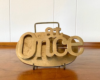 May include: A brass sign with the words "at once" in a stylized font. The sign is on a metal stand.