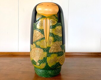 May include: A wooden Japanese doll with a green and gold patterned kimono. The doll has black hair and a closed-eye expression.