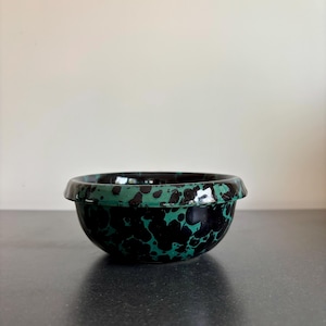 May include: A small, round bowl with a black and green speckled pattern. The bowl has a slightly flared rim and a glossy finish. The interior is black, and the exterior features a dark green base with black spots. The bowl is likely ceramic or porcelain.