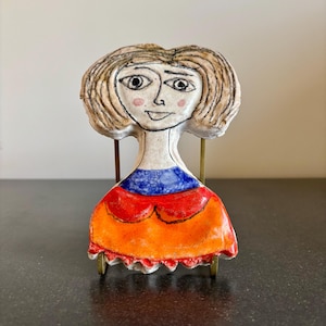 May include: A ceramic figurine of a woman with a whimsical design. The figure has a cream-colored face with pink cheeks, black eyes, and a small smile. The dress is orange and red with a blue neckline. The figurine is mounted on a brass stand.