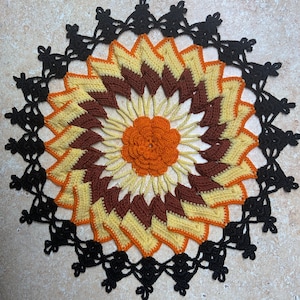 May include: A crocheted doily with a black lace border and a multi-colored center featuring shades of orange, yellow, and brown.