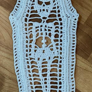 May include: White crochet coffin-shaped doily. The openwork design features a skull and floral motifs. The edges are scalloped. The doily is placed on a wooden surface.